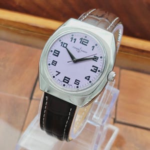 Puede incluir: Un reloj de pulsera plateado con esfera lila claro y n&uacute;meros negros. El reloj tiene una correa de cuero marr&oacute;n con costuras blancas. La esfera muestra la marca "Ulysse Nardin" y "Swiss Made".