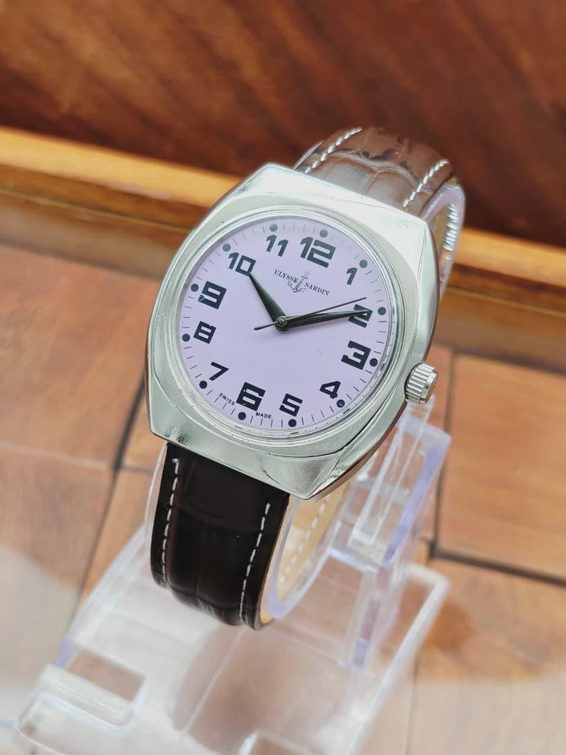 Puede incluir: Un reloj de pulsera plateado con esfera lila claro y n&uacute;meros negros. El reloj tiene una correa de cuero marr&oacute;n con costuras blancas. La esfera muestra la marca "Ulysse Nardin" y "Swiss Made".