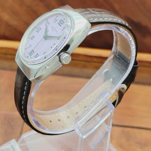 Puede incluir: Un reloj de pulsera plateado con esfera lila claro y correa de cuero negro. El reloj tiene una caja rectangular y una correa texturizada. La esfera del reloj presenta n&uacute;meros y agujas negros. El reloj se exhibe sobre un soporte de pl&aacute;stico transparente.