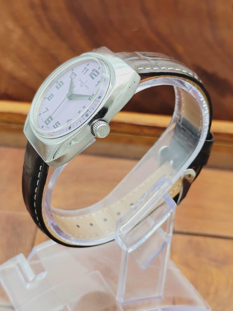 Puede incluir: Un reloj de pulsera plateado con esfera lila claro y correa de cuero negro. El reloj tiene una caja rectangular y una correa texturizada. La esfera del reloj presenta n&uacute;meros y agujas negros. El reloj se exhibe sobre un soporte de pl&aacute;stico transparente.