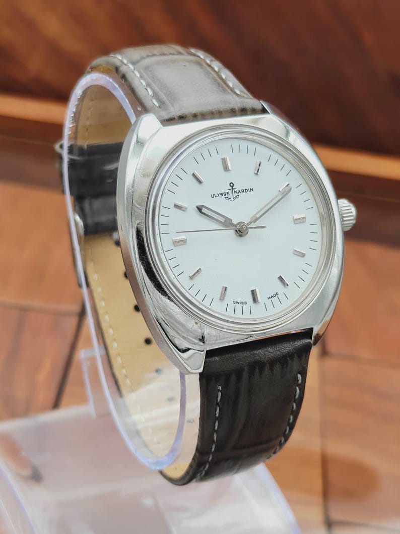 May include: A silver-toned wristwatch with a white face and black leather band. The watch features the brand name "Ulysse Nardin" and has a classic design. The band is approximately 8 inches long.