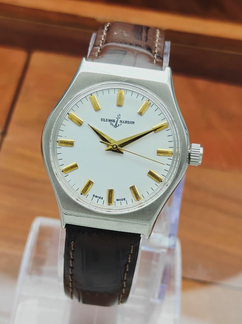 May include: A silver Ulysse Nardin wristwatch with a white face and gold hands and markers. The watch has a brown leather strap. The watch face displays the text "Ulysse Nardin" and "Swiss Made".