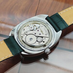 K&ouml;nnte beinhalten: Nahaufnahme des Uhrwerks einer Vintage-Armbanduhr. Das silberfarbene Geh&auml;use zeigt die komplizierten Zahnr&auml;der und Komponenten. Es hat ein gr&uuml;nes Lederarmband und eine Krone. Das Innenleben der Uhr ist sichtbar und unterstreicht die Handwerkskunst.
