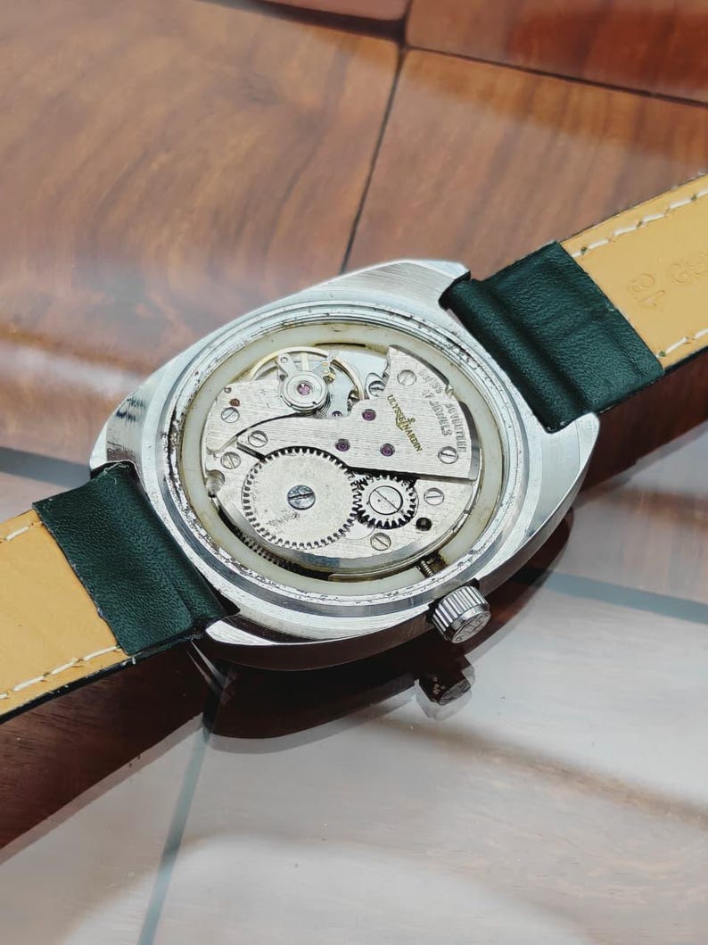 K&ouml;nnte beinhalten: Nahaufnahme des Uhrwerks einer Vintage-Armbanduhr. Das silberfarbene Geh&auml;use zeigt die komplizierten Zahnr&auml;der und Komponenten. Es hat ein gr&uuml;nes Lederarmband und eine Krone. Das Innenleben der Uhr ist sichtbar und unterstreicht die Handwerkskunst.