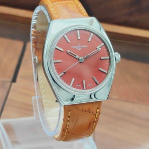 Puede incluir: Un reloj de pulsera Ulysse Nardin vintage con esfera roja y caja plateada. El reloj tiene una correa de cuero marr&oacute;n con un patr&oacute;n de cocodrilo. La esfera del reloj tiene marcadores de hora y agujas plateadas.