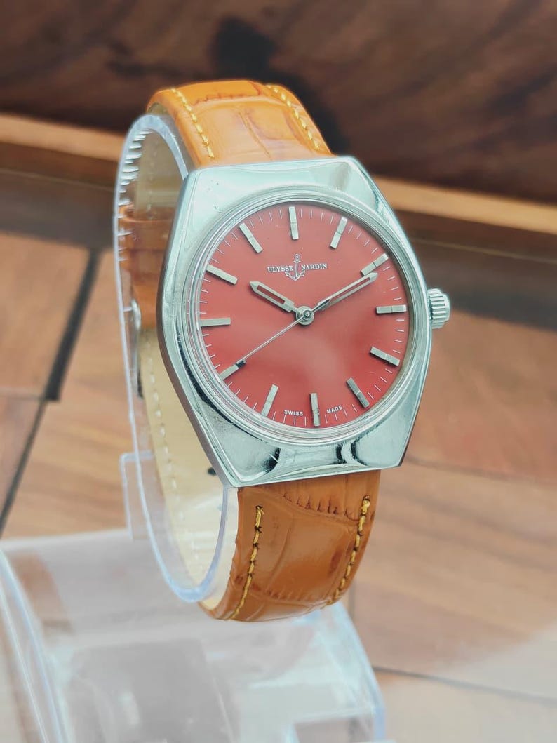 Puede incluir: Un reloj de pulsera Ulysse Nardin vintage con esfera roja y caja plateada. El reloj tiene una correa de cuero marr&oacute;n con un patr&oacute;n de cocodrilo. La esfera del reloj tiene marcadores de hora y agujas plateadas.