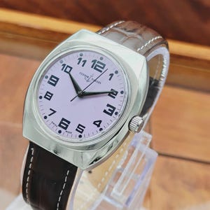 Puede incluir: Un reloj de pulsera plateado con esfera lila claro y n&uacute;meros negros. El reloj tiene una correa de cuero marr&oacute;n con costuras blancas. La esfera del reloj dice "Ulysse Nardin" y "Swiss Made".