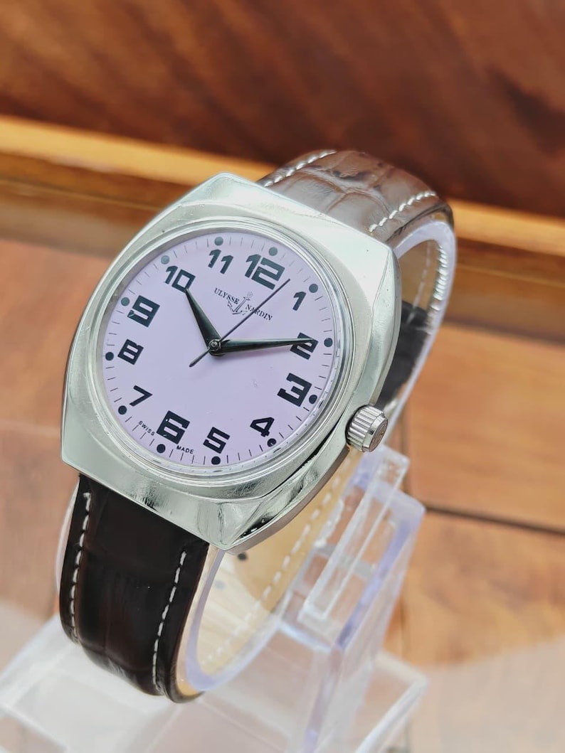 Puede incluir: Un reloj de pulsera plateado con esfera lila claro y n&uacute;meros negros. El reloj tiene una correa de cuero marr&oacute;n con costuras blancas. La esfera del reloj dice "Ulysse Nardin" y "Swiss Made".