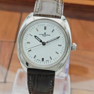May include: A silver-toned wristwatch with a white face and black hands. The watch has a brown leather band and the brand name "ULYSSE NARDIN" is visible on the face. The watch is marked "SWISS MADE".