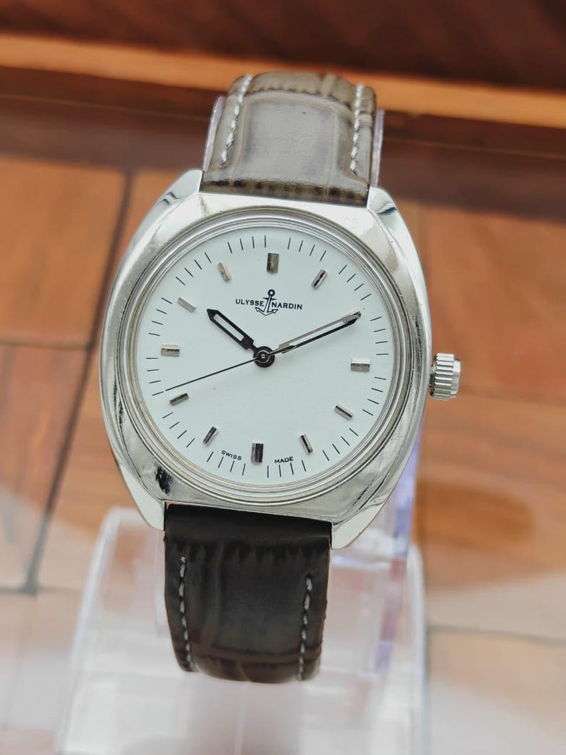 May include: A silver-toned wristwatch with a white face and black hands. The watch has a brown leather band and the brand name "ULYSSE NARDIN" is visible on the face. The watch is marked "SWISS MADE".