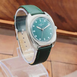 K&ouml;nnte beinhalten: Eine silberfarbene Armbanduhr mit gr&uuml;nem Zifferblatt und gr&uuml;nem Lederarmband. Die Uhr hat ein rundes Zifferblatt mit silbernen Zeigern und Stundenmarkierungen. Das Armband ist mit gr&uuml;nem Faden gen&auml;ht. Die Uhr wird auf einem transparenten Kunststoffst&auml;nder ausgestellt.