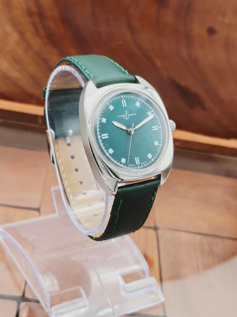 K&ouml;nnte beinhalten: Eine silberfarbene Armbanduhr mit gr&uuml;nem Zifferblatt und gr&uuml;nem Lederarmband. Die Uhr hat ein rundes Zifferblatt mit silbernen Zeigern und Stundenmarkierungen. Das Armband ist mit gr&uuml;nem Faden gen&auml;ht. Die Uhr wird auf einem transparenten Kunststoffst&auml;nder ausgestellt.