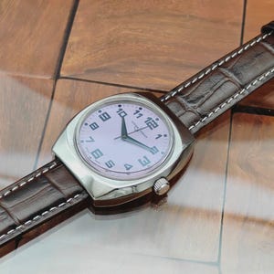 Puede incluir: Un reloj de pulsera plateado con esfera rosa claro y n&uacute;meros negros. El reloj tiene una correa de cuero marr&oacute;n con costuras blancas. La esfera muestra la marca "LEONARD HARDIN".