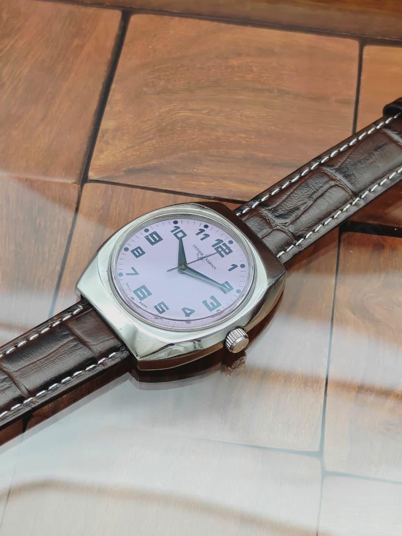 Puede incluir: Un reloj de pulsera plateado con esfera rosa claro y n&uacute;meros negros. El reloj tiene una correa de cuero marr&oacute;n con costuras blancas. La esfera muestra la marca "LEONARD HARDIN".