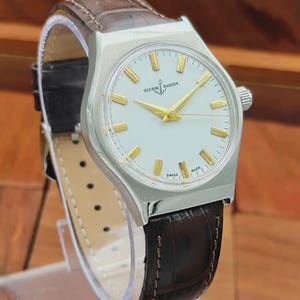 May include: A vintage Ulysse Nardin wristwatch with a silver case and a white dial. The watch has gold hands and markers, and a brown leather strap. The watch is displayed on a clear stand.