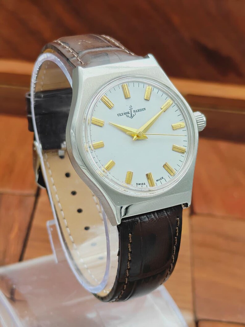 May include: A vintage Ulysse Nardin wristwatch with a silver case and a white dial. The watch has gold hands and markers, and a brown leather strap. The watch is displayed on a clear stand.