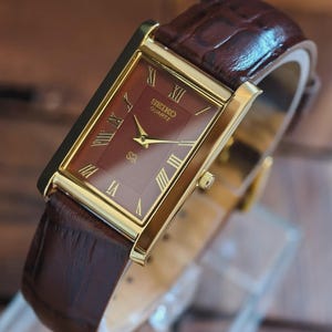 Puede incluir: Una taza de cerámica blanca con un asa negra y el texto "But First, Coffee" en negrita negra. La taza se encuentra sobre un fondo neutro, resaltando su diseño sencillo.