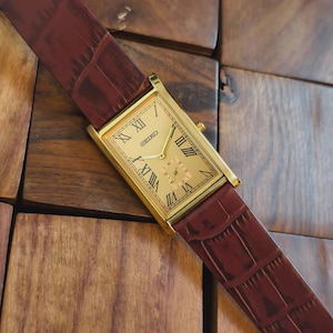 May include: A gold-toned rectangular wristwatch with a cream-colored face and Roman numeral hour markers. The watch features the brand name "SEIKO" and is set on a burgundy leather strap with a crocodile pattern.