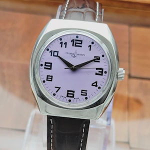 Puede incluir: Un reloj de pulsera Ulysse Nardin plateado con esfera lila claro y agujas negras. El reloj tiene una correa de cuero marr&oacute;n con costuras blancas. La esfera muestra los n&uacute;meros del 1 al 12.