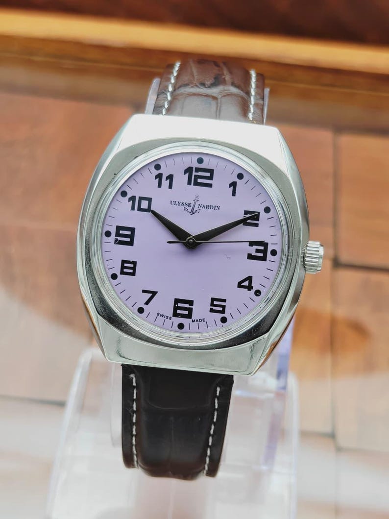 Puede incluir: Un reloj de pulsera Ulysse Nardin plateado con esfera lila claro y agujas negras. El reloj tiene una correa de cuero marr&oacute;n con costuras blancas. La esfera muestra los n&uacute;meros del 1 al 12.