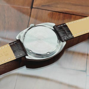 Puede incluir: Un reloj de pulsera plateado con una correa de cuero marr&oacute;n. La caja del reloj es redonda con una parte trasera texturizada. La correa es de color marr&oacute;n oscuro con una parte inferior beige claro y costuras blancas. El reloj est&aacute; sobre una superficie de madera.