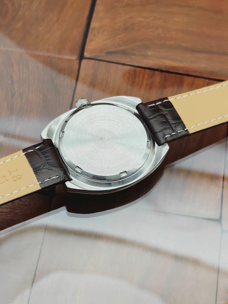 Puede incluir: Un reloj de pulsera plateado con una correa de cuero marr&oacute;n. La caja del reloj es redonda con una parte trasera texturizada. La correa es de color marr&oacute;n oscuro con una parte inferior beige claro y costuras blancas. El reloj est&aacute; sobre una superficie de madera.