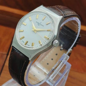 May include: A silver Ulysse Nardin wristwatch with a light-coloured face and gold hands and markers. The watch has a brown leather strap. The watch is displayed on a clear plastic stand.