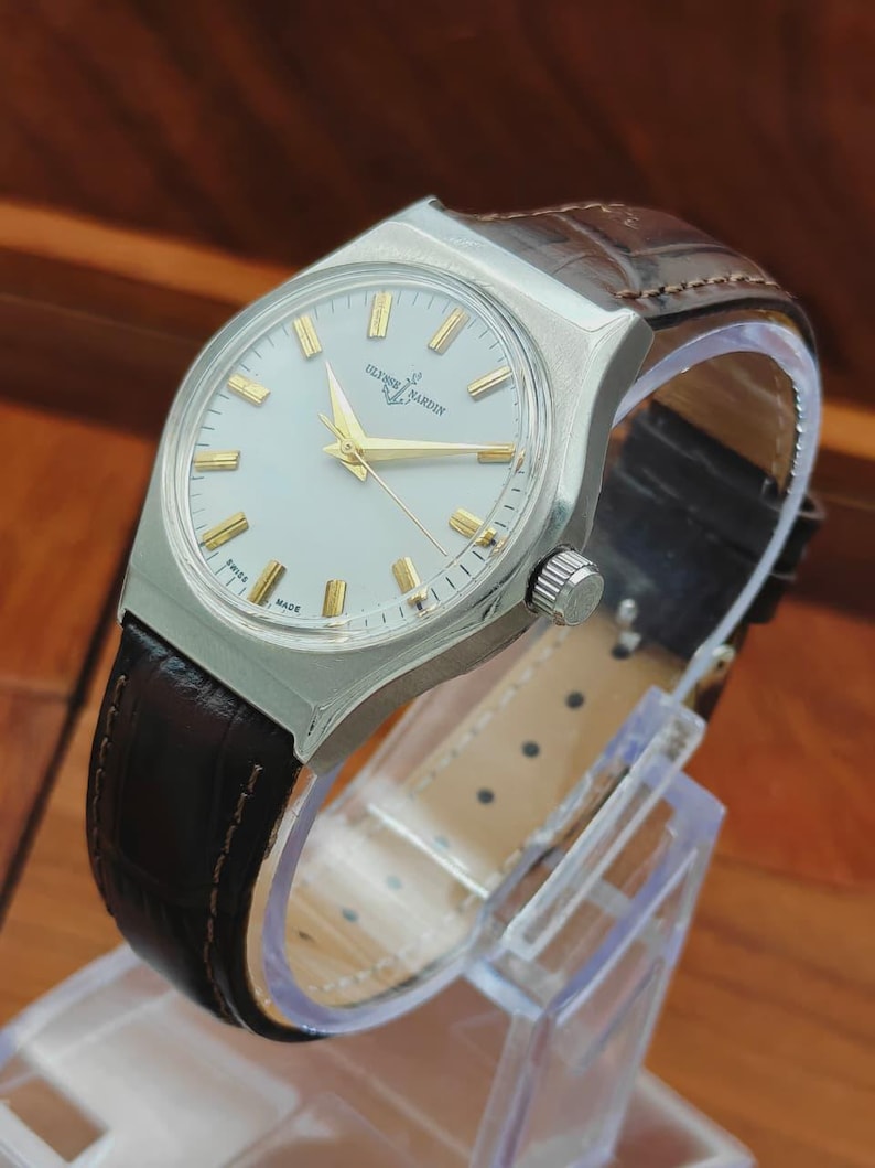 May include: A silver Ulysse Nardin wristwatch with a light-coloured face and gold hands and markers. The watch has a brown leather strap. The watch is displayed on a clear plastic stand.