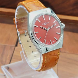 Puede incluir: Un reloj Ulysse Nardin vintage con esfera roja y caja plateada. La correa es de cuero marr&oacute;n con un patr&oacute;n de cocodrilo. El reloj se exhibe sobre un soporte transparente.