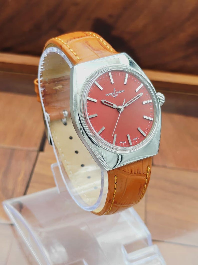Puede incluir: Un reloj Ulysse Nardin vintage con esfera roja y caja plateada. La correa es de cuero marr&oacute;n con un patr&oacute;n de cocodrilo. El reloj se exhibe sobre un soporte transparente.
