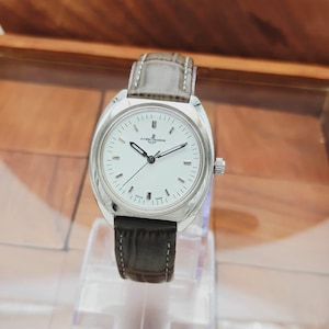 May include: A silver-toned wristwatch with a white face and a dark gray leather band. The watch has a rectangular face with black hands and hour markers. The watch is displayed on a clear stand.