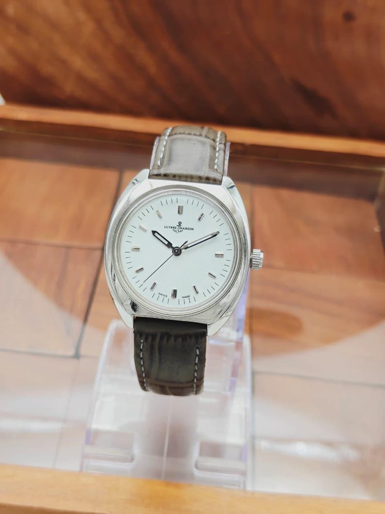 May include: A silver-toned wristwatch with a white face and a dark gray leather band. The watch has a rectangular face with black hands and hour markers. The watch is displayed on a clear stand.