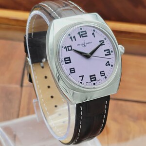 Puede incluir: Un reloj de pulsera Ulysse Nardin plateado con esfera lila claro y agujas negras. El reloj tiene una correa de cuero marr&oacute;n con costuras blancas. La esfera muestra n&uacute;meros negros y las palabras "Ulysse Nardin" y "Swiss Made".
