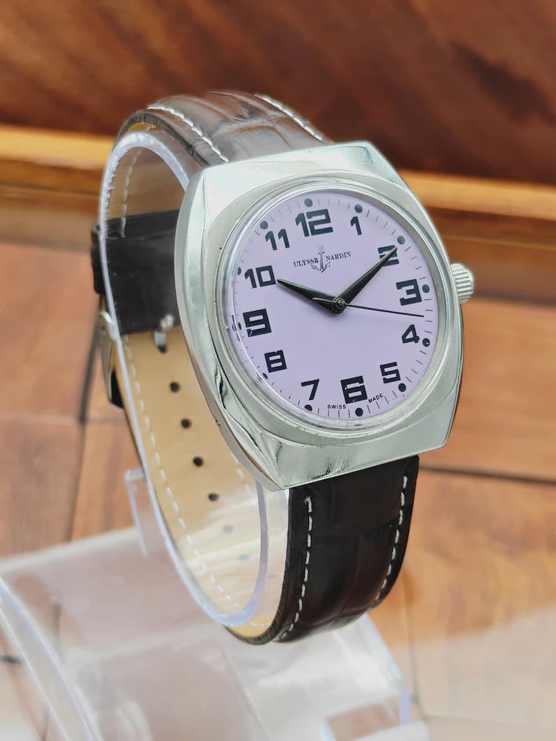 Puede incluir: Un reloj de pulsera Ulysse Nardin plateado con esfera lila claro y agujas negras. El reloj tiene una correa de cuero marr&oacute;n con costuras blancas. La esfera muestra n&uacute;meros negros y las palabras "Ulysse Nardin" y "Swiss Made".