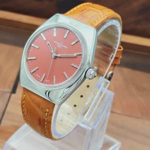 Puede incluir: Un reloj Ulysse Nardin vintage con una caja cuadrada plateada y una esfera color coral. El reloj presenta agujas e indicadores plateados y una correa de cuero marr&oacute;n. El reloj se exhibe sobre un soporte transparente.