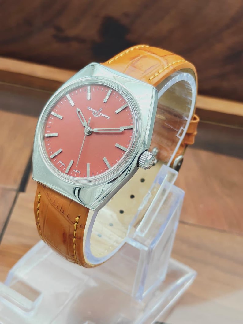 Puede incluir: Un reloj Ulysse Nardin vintage con una caja cuadrada plateada y una esfera color coral. El reloj presenta agujas e indicadores plateados y una correa de cuero marr&oacute;n. El reloj se exhibe sobre un soporte transparente.