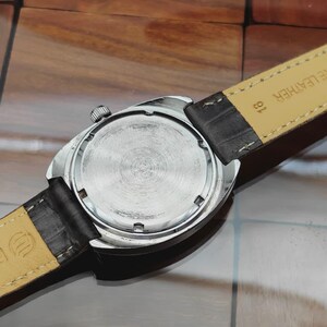 May include: Close-up of a vintage wristwatch, showcasing the watch's back and leather strap. The watch has a silver-colored case and a textured, circular back. The black leather strap is attached to the watch case.