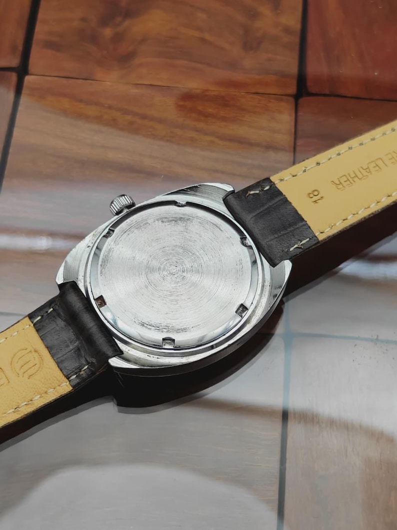 May include: Close-up of a vintage wristwatch, showcasing the watch's back and leather strap. The watch has a silver-colored case and a textured, circular back. The black leather strap is attached to the watch case.