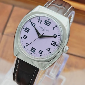 Puede incluir: Un reloj de pulsera Ulysse Nardin plateado con esfera lila claro y n&uacute;meros y agujas negras. El reloj tiene una correa de cuero marr&oacute;n. La esfera muestra el nombre de la marca y "SWISS MADE".