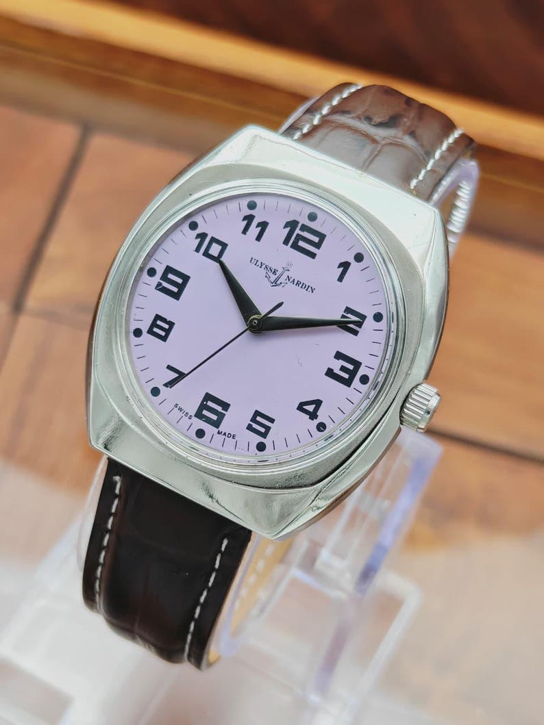 Puede incluir: Un reloj de pulsera Ulysse Nardin plateado con esfera lila claro y n&uacute;meros y agujas negras. El reloj tiene una correa de cuero marr&oacute;n. La esfera muestra el nombre de la marca y "SWISS MADE".