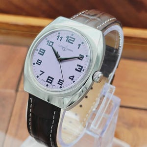 Puede incluir: Un reloj de pulsera Ulysse Nardin plateado con esfera rosa claro y n&uacute;meros negros. El reloj tiene una correa de cuero marr&oacute;n con costuras blancas. El reloj se exhibe sobre un soporte transparente.