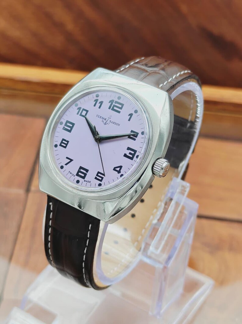 Puede incluir: Un reloj de pulsera Ulysse Nardin plateado con esfera rosa claro y n&uacute;meros negros. El reloj tiene una correa de cuero marr&oacute;n con costuras blancas. El reloj se exhibe sobre un soporte transparente.