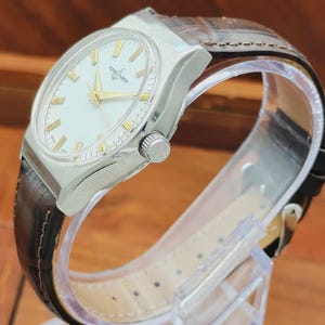 May include: A silver wristwatch with a white face and gold hands and markers. The watch has a brown leather strap. The watch is displayed on a clear plastic stand.