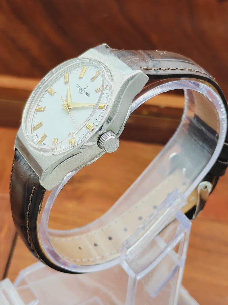 May include: A silver wristwatch with a white face and gold hands and markers. The watch has a brown leather strap. The watch is displayed on a clear plastic stand.