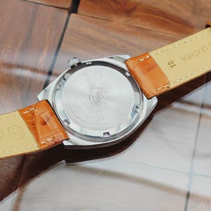 Puede incluir: Un reloj de pulsera plateado con una correa de cuero marr&oacute;n. La parte trasera de la caja del reloj es visible, mostrando un dise&ntilde;o circular de metal cepillado. La correa es de color marr&oacute;n claro con un patr&oacute;n texturizado. El reloj tiene una corona plateada.