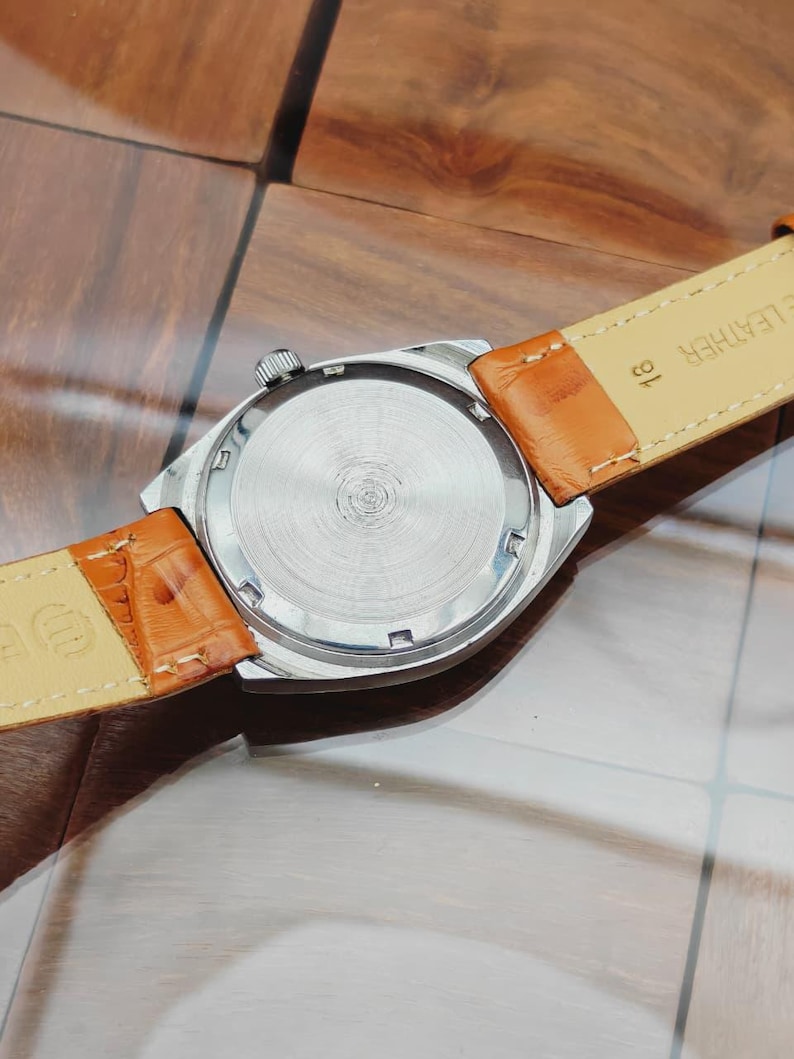 Puede incluir: Un reloj de pulsera plateado con una correa de cuero marr&oacute;n. La parte trasera de la caja del reloj es visible, mostrando un dise&ntilde;o circular de metal cepillado. La correa es de color marr&oacute;n claro con un patr&oacute;n texturizado. El reloj tiene una corona plateada.
