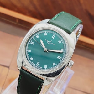 May include: A vintage Ulysse Nardin wristwatch with a green dial and a green leather strap. The watch has a silver-colored case and a date window. The watch is displayed on a clear stand.