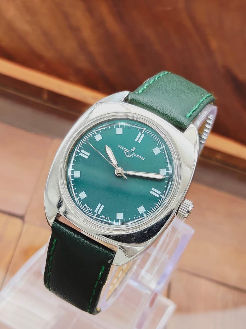 K&ouml;nnte beinhalten: Eine Vintage-Armbanduhr von Ulysse Nardin mit gr&uuml;nem Zifferblatt und gr&uuml;nem Lederarmband. Die Uhr hat ein silberfarbenes Geh&auml;use und ein Datumsfenster. Die Uhr wird auf einem transparenten St&auml;nder ausgestellt.