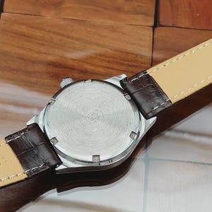 May include: A silver wristwatch with a brown leather strap. The watch face is not visible. The back of the watch has a circular, textured design. The watch strap is brown with a light beige underside.