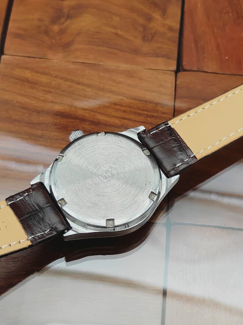 May include: A silver wristwatch with a brown leather strap. The watch face is not visible. The back of the watch has a circular, textured design. The watch strap is brown with a light beige underside.